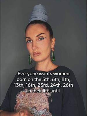 A head and shoulders shot of a woman looking at the camera. She has light skin, dark eyeliner, and plum lipstick. She has a gray, turban-like head covering and large, green and gold earrings. A black t-shirt showing some kind of brown picture appears between her neck and the text at the bottom of the image.