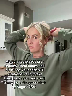 A woman with blonde hair pulled back, wearing a green sweater, is looking towards the camera while holding the sides of her hair as if putting it up. In the background is a kitchen.