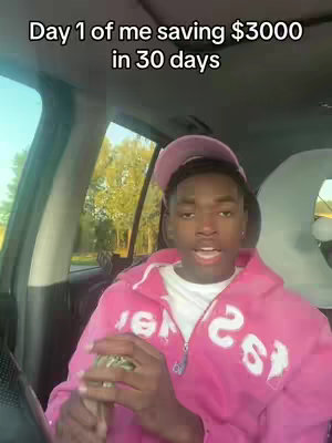 A young man in a pink hoodie and cap sits inside a car, speaking to the camera. Text overlayed on the video reads, 'Day 1 of me saving $3000 in 30 days.' The background subtly reveals trees, suggesting an outdoor setting.
