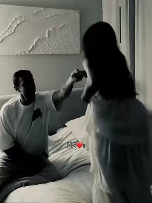 A black and white image shows a man and a woman in a bedroom. The man is sitting on the bed, wearing a t-shirt, and holding the woman's hand. The woman is standing, wearing a light-colored dress. The bed is neatly made with white sheets and pillows. A textured artwork hangs on the wall above the bed. The overall composition is intimate and romantic.