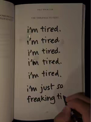 The image captures an overhead view of an open notebook on a dark surface. The page is filled with the handwritten phrase "i'm tired" repeated multiple times. A hand holding a black pen is visible, with the pen's nib near the bottom right, as if writing the last words. The overall color scheme leans toward a dim, low-light setting.