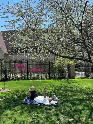A woman wearing a hat and white outfit is lying on a blue blanket on a green lawn. Her legs are up, and there's a tree with white blossoms overhead. A black wrought-iron fence and a building are in the background.