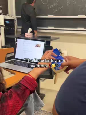 A candid shot from a classroom. A person in a red plaid shirt is holding a blue item open, while the hand of another person in a blue shirt is opening the item. A laptop displaying a webpage is open nearby, to the left. In the background, a teacher stands facing a chalkboard.