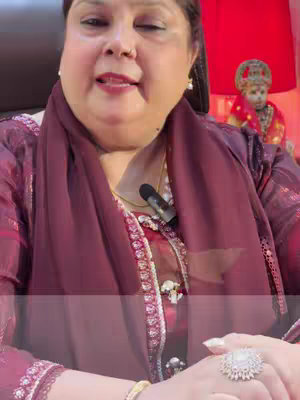 A woman with a dark red shawl and crimson embroidered dress is addressing the camera directly. A microphone hangs near her collarbone, and she wears a large ring with a white sparkling stone on one hand. Behind her is a bright red light.