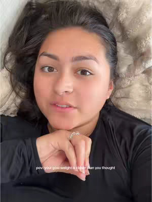 A woman is shown from a close, slightly above, angle, lying down. She looks directly at the camera with a neutral expression. She appears to be lying on a soft, textured surface that is light-colored. Her dark hair is spread out behind her. She wears a black top and has a ring on her finger. The text 'pov: your goal weight is closer than you thought' is overlaid at the bottom of the image.