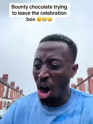 A man with short dark hair and a blue shirt is shown from the chest up, with the background in a blurred gray, cloudy outdoor setting.