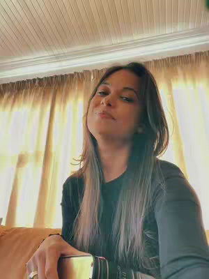 A woman with long, highlighted hair looks up at the camera with a slight smile. She is seated indoors, with a partly obscured background of curtains and a window. She is holding a phone or similar device in her hands.