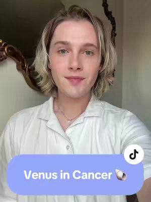 A selfie-style image featuring a young man with fair skin and blonde hair, posing in front of a blurred background. He is wearing a collared white shirt and a delicate silver necklace. A light-blue, oval-shaped text overlay at the bottom of the frame reads 'Venus in Cancer' with a seashell icon next to it. The man is smiling slightly and looking directly at the camera.