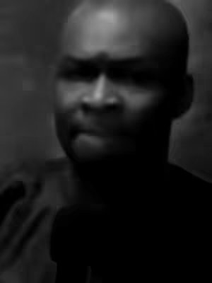 A monochrome image of a man facing the camera, with a serious expression. His face is the primary focus, with a dark background. The lighting emphasizes the facial features.