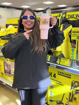 A woman with sunglasses smiles, holding a light pink tumbler in a retail setting. The background features store displays with yellow and black product packaging.