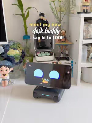 A well-lit desk setup with a white surface. The focal point is a small, dark gray robot with a screen displaying blue cartoon features, positioned in the center. Behind the robot are various accessories and decorations, including a small figure of a person, a plant. Text overlays at the top, in a sans-serif font, reads 'meet my new desk buddy - say hi to LOOI!!' The overall composition is tidy.
