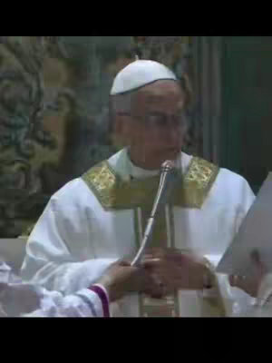 The cover image shows a medium shot of a man, likely a religious figure, standing and speaking. He is wearing a white garment with gold and white accents around the shoulders and neck and a white skullcap. He holds a microphone with his left hand and appears to be reading from papers held in his right hand. The background is out of focus, suggesting an interior setting with hints of architectural details and other people.