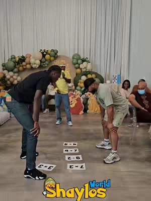 Two men are bent over, looking at pieces of paper with math problems listed on them. A man looks at the people playing from an elevated surface. Decorations consist of balloons and Lion King cutouts.