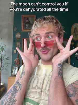 A man with blonde hair and pink-tinted glasses looks at the camera with a surprised expression, his hands up near his face. The background is a dark teal wall adorned with multiple framed pictures. He wears a yellow shirt and has tattoos on both arms. The hook says: "The moon can't control you if you're dehydrated all the time".