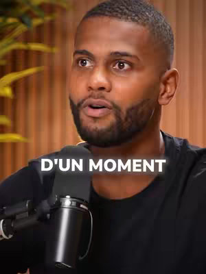 A medium shot features a Black man with short, dark hair and a well-groomed beard. He is wearing a black t-shirt and holding a microphone close to his mouth. The text overlay reads, "D'UN MOMENT". The background is blurred, suggesting a studio or podcast setting.