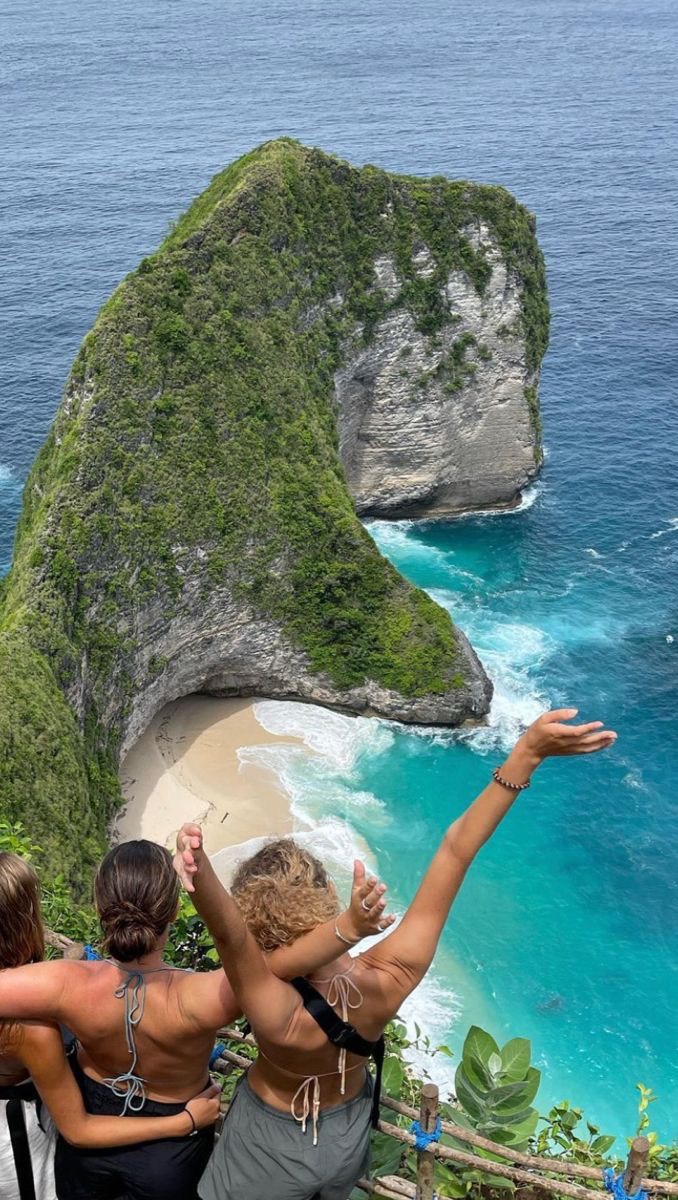 An overhead shot captures three women looking out over a scenic coastal vista. The frame features a huge rock formation covered in vibrant green foliage, jutting out into the turquoise ocean. The women are seen from the back, with two arms raised, amplifying the sense of awe and appreciation of the beautiful setting. The composition is balanced with warm tones, emphasizing the natural beauty of the ocean and landscape.