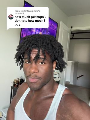 A selfie-style shot featuring a muscular Black man. He has short, dark, dreadlocked hair and a trimmed mustache. He's wearing a white tank top, with the upper body in full view. Behind him is a somewhat cluttered room with a door and items visible on a shelf.