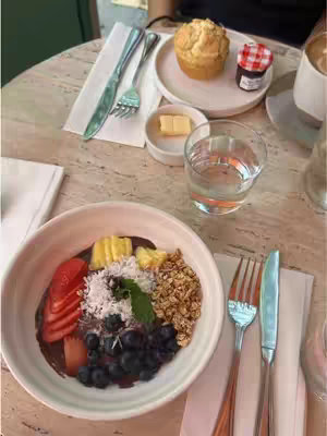 The image shows a table setting with brunch items. A large white bowl with a colorful arrangement of sliced strawberries, blueberries, coconut flakes, granola, and a sprig of mint sits at the front. Adjacent, a plate holds a muffin and small dish of jam next to a glass of water and a coffee cup. Silverware and a napkin are also visible.