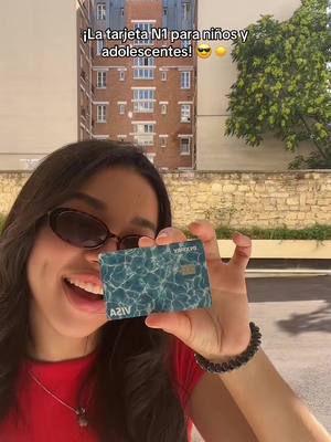 A woman smiling and holding up a turquoise and white credit card. She is wearing sunglasses and a red top. There is a building in the background.