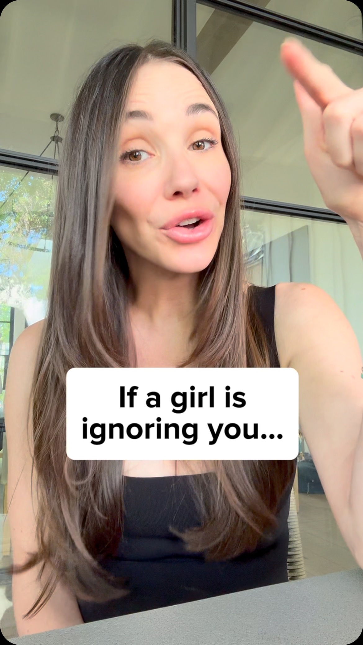 A woman with long brown hair is shown in a selfie-style shot. She is looking directly at the camera with her mouth open as if she is speaking. She is wearing a black top. A white rectangle with black text covers a portion of the bottom of her body. She is pointing upward with her right hand. Behind her is a window that is partially visible. There is a pendant light.