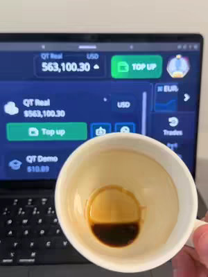 The image features a laptop screen displaying a financial platform, with a cup of coffee in the foreground. The screen shows account details with a high balance of $563,100.30. A semi-filled coffee cup dominates the foreground, partially obscuring the laptop. The cup's interior shows a dark coffee stain in the bottom half.
