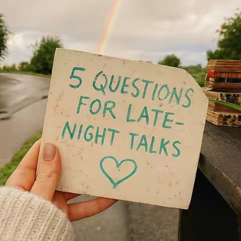 The image shows a hand holding a square, slightly damaged piece of paper. On the paper, there is handwritten text in teal-colored marker that reads "5 QUESTIONS FOR LATE-NIGHT TALKS" with a heart symbol drawn below the text. The background is soft, with a rainbow and an overcast sky, along with greenery and a road in the distance. The overall composition has a warm tone, possibly capturing a moment outdoors.