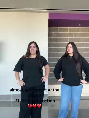Two young women stand side-by-side in what appears to be a hallway, facing the camera. The woman on the left is dressed in a black t-shirt and black pants, while the woman on the right is wearing a black long-sleeved shirt and light blue jeans. The background includes a wall with a section of purple. Text overlaid on the image reads 'almost got caught w the phone plans #ihateiphoneban' with a series of hearts.