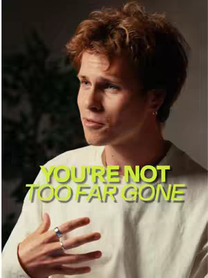 A young man with fair skin and short, curly auburn hair is centered in the frame. He is wearing a white long-sleeved shirt and has a silver ring on his left hand. The text overlay says: You’re not too far gone. The background is blurred but appears to be an interior setting.
