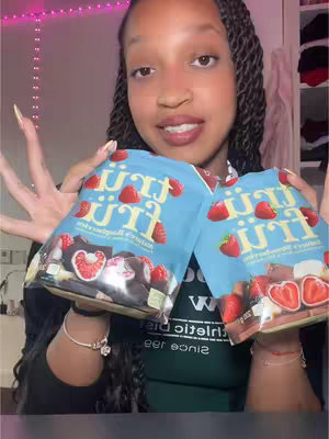A young Black woman with long braided hair is holding two packages of Trüfrü chocolate-covered strawberries in front of a light-pink-lit room. She is smiling and looking at the camera. The Trüfrü packages are light blue with images of chocolate-covered strawberries on them, and she wears a black top along with a green shirt. There is a white closet in the background, full of clothing, and she is wearing bracelets. Her nails are long and acrylic with nail polish.