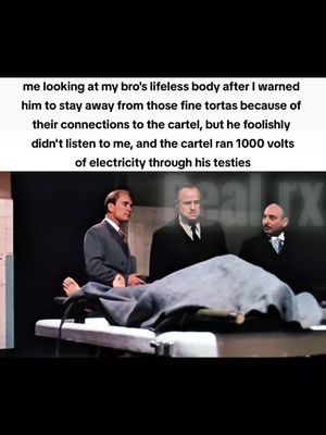 A still image from the film "The Godfather". The image is framed vertically and depicts a somber scene in what appears to be a morgue or sterile room. Three men stand around a table, where a body is laid out, covered by a sheet. One man in the foreground is examining the body. The color palette is muted, with dark clothing and a gray background, creating a serious, dramatic tone.