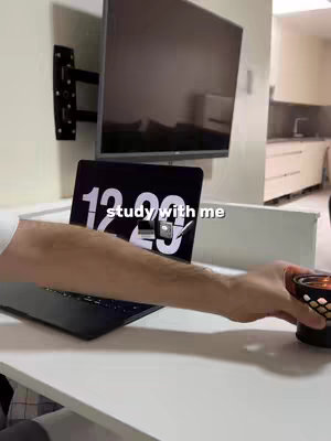 A person's arm reaching towards a lit candle on a white desk, with a laptop showing the time. A large TV is mounted on the wall in the background, above some kitchen cabinets.