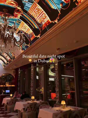 An interior shot of a luxurious restaurant. A large, ornate chandelier hangs from the ceiling. The ceiling itself is decorated with a colorful, artistic design. Tables are set with white tablecloths and simple lamps. The windows show outside lights.