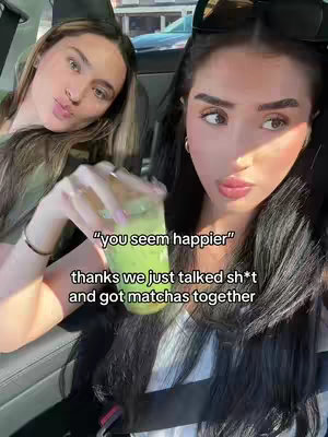 Two women are seated inside a car, taking a selfie. The woman on the right holds a matcha drink. Overlayed text reads, "you seem happier" and "thanks we just talked sh*t and got matchas together."