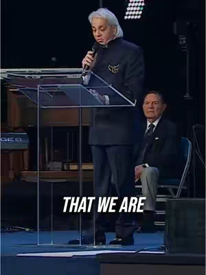 A man in a black suit with a microphone stands behind a clear podium, speaking. Another man is seated to his right wearing a suit and tie. The background is a dark stage setup.