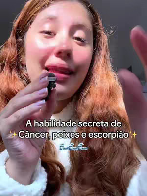 A young woman with long, wavy reddish-brown hair is holding a small microphone close to her lips. The background is softly blurred, and the woman appears to be speaking or about to speak. A text overlay at the bottom of the image says, 'A habilidade secreta de Câncer, peixes e escorpião' with added sparkles and a watery design.