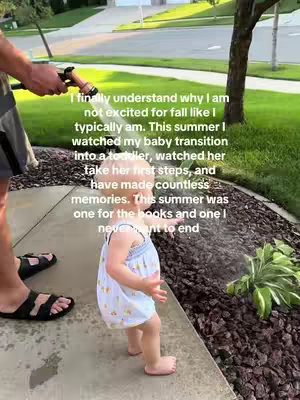 A toddler in a light blue dress with a floral pattern stands on a stone walkway, facing a stream of water. An arm holding a hose directs the water towards the child. The background includes a green lawn, a tree, and a glimpse of a residential street with houses.