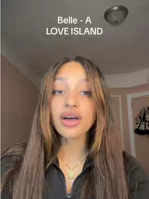 A selfie-style shot of a young woman with long brown hair. The woman is looking towards the camera with her mouth slightly open. There is text overlay at the top of the image with the labels "Belle - A LOVE ISLAND". The background is out of focus and includes what appears to be a light fixture.