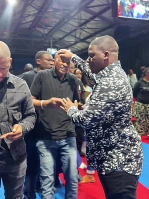 A man is being prayed for in a church setting. The main focus is on two men; one has his hand on the other's head. Other people are in the background watching. The scene is brightly lit.
