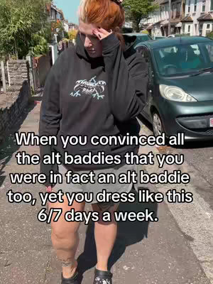 A person wearing a black hoodie and gray shorts stands outdoors on a sunny day. The person has red-orange hair and is looking down. Text on the image describes their clothing choices.