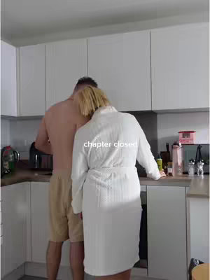 A couple is in a kitchen. The man is shirtless with khaki shorts, appearing to be cooking while the woman, with blonde hair, is in a white robe leaning against him. White cabinets form the backdrop.