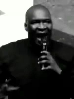 A grayscale video still shows a man in a black shirt holding a microphone and speaking. The background is out of focus. The man has short hair and is slightly smiling.