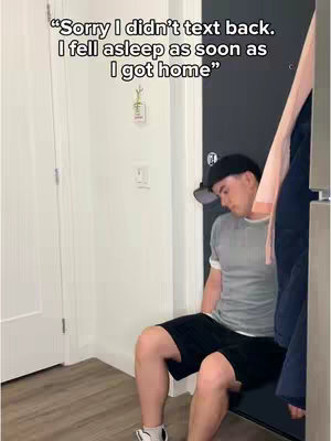 A man in a gray t-shirt, black shorts, and a black baseball cap is sitting hunched over on a bench along a white wall. Text is overlaid on the image. He looks tired or sleepy.