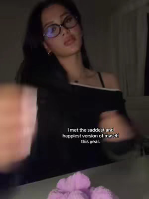The cover image shows a woman wearing glasses, looking slightly down with a thoughtful expression. The background is dimly lit. In the foreground, there is an out-of-focus hand. At the bottom, a pile of purple objects is visible. The overlay text states, "i met the saddest and happiest version of myself this year."