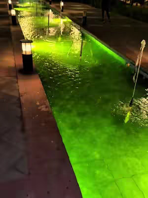 A nighttime shot of a modern garden with a reflective water feature. A channel of flowing water, lit with a bright green hue, is the central focus. Small lights line the edges of the water, and there's a paved area with a person strolling in the background. The overall ambiance is serene and well-lit.
