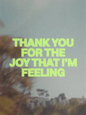 A vertical image featuring a blurry background of trees and a light blue sky. Overlaid on this is bold, neon green text that reads, "THANK YOU FOR THE JOY THAT I'M FEELING."
