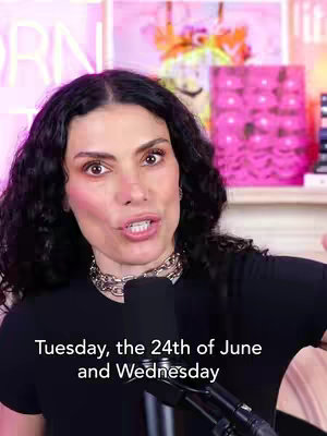 A woman with dark curly hair and a black shirt is in front of a microphone. Behind her is a shelf with items, including a framed piece of art lit with pink lights. Text at the bottom of the screen displays the day of the week.