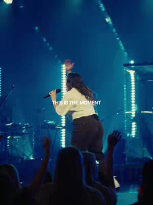 A woman with long dark hair sings into a microphone on a stage bathed in blue light. The text 'THIS IS THE MOMENT' is overlaid in white toward the bottom left. The audience is blurred in the foreground.