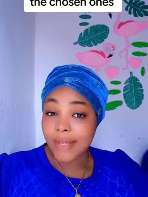 A woman with a blue headscarf and shirt is looking at the camera. In the background, a wall with pink flamingo and green leaf decals can be seen.