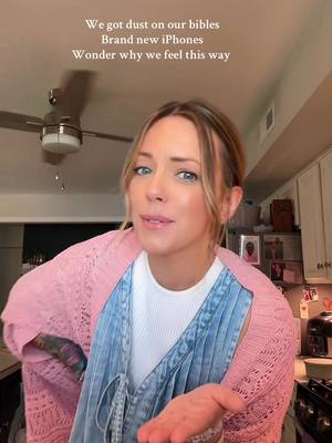 A woman in a pink cardigan and denim overalls is shown with a slight smile, hands outstretched. Text overlays read, 'We got dust on our bibles, Brand new iPhones, Wonder why we feel this way.' The upper portion of the image shows a ceiling with a fan.