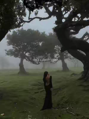 A woman in a long black dress stands in a misty forest clearing, framed by large, shadowy trees. The scene is bathed in a soft green light, creating a moody and atmospheric feel. The text overlay reads "she's a earth sign".